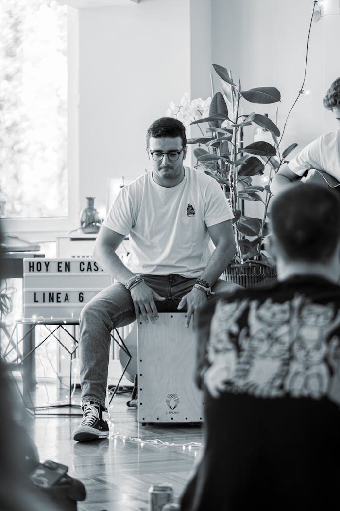 A black and white photo of a young man playing the cajon during an indoor jam session.